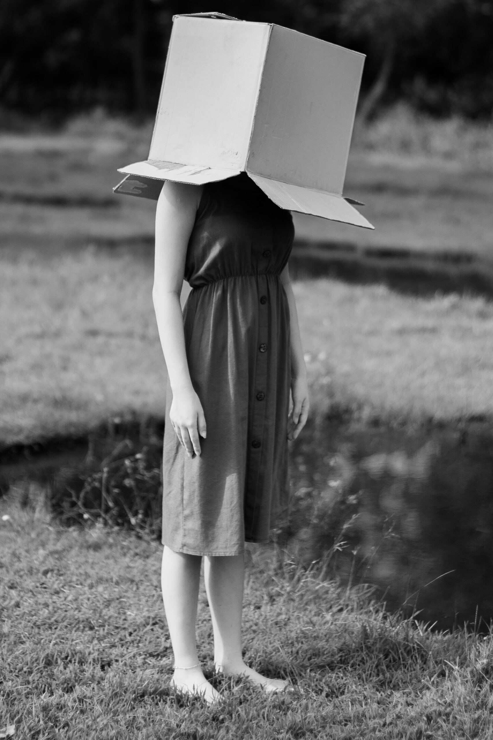 Black and white photo of a woman outdoors in nature, head hidden in a box.